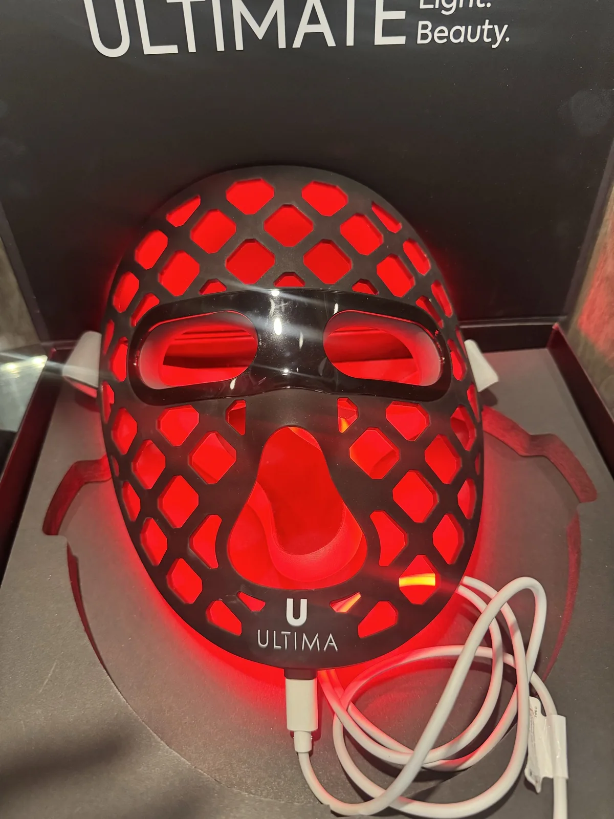 Az ULTIMA LED Beauty Mask bekapcsolva, vörös fénnyel, a saját dobozában